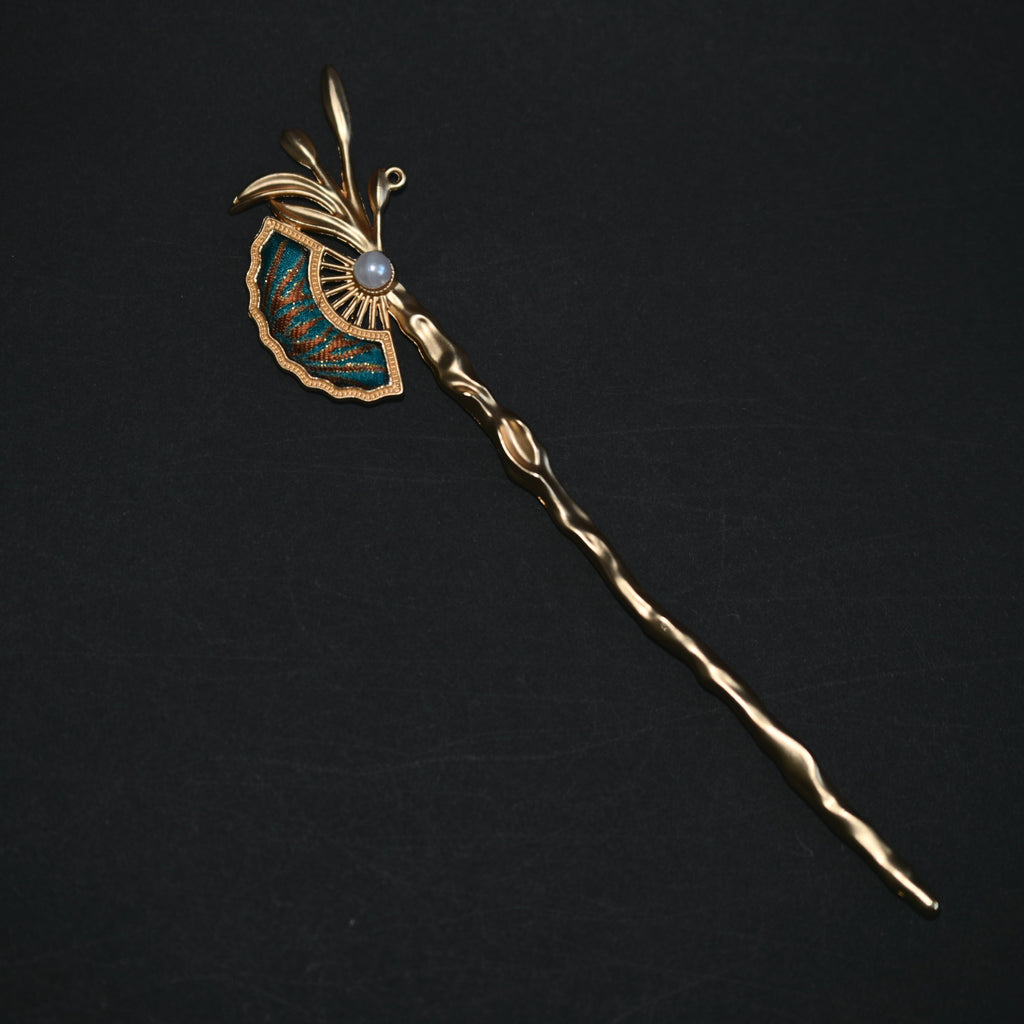 A vintage gold hairpin with a decorative fan-shaped enamel inlay, a central pearl-like bead, and a long twisted stem, displayed on a dark background.