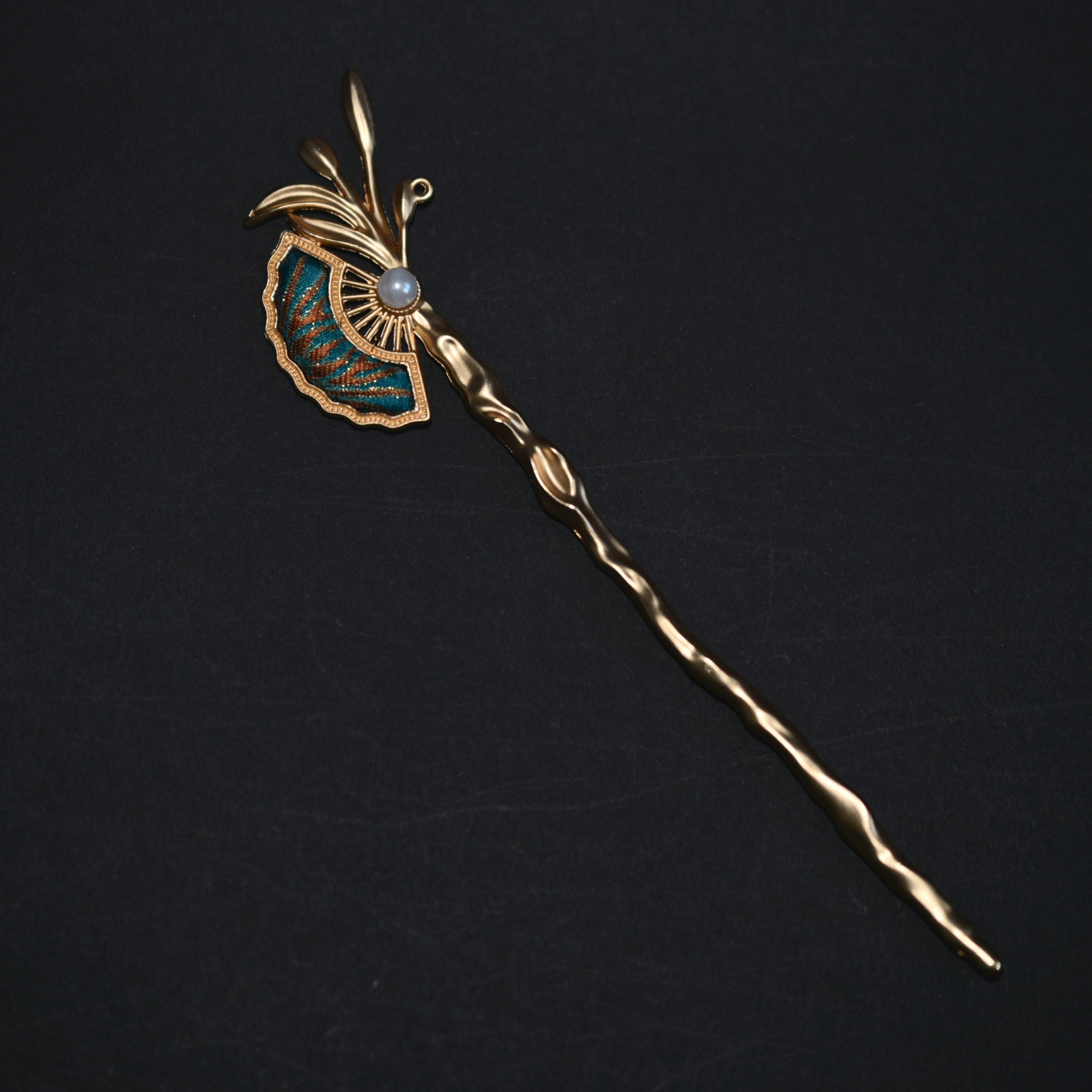 A vintage gold hairpin with a decorative fan-shaped enamel inlay, a central pearl-like bead, and a long twisted stem, displayed on a dark background.